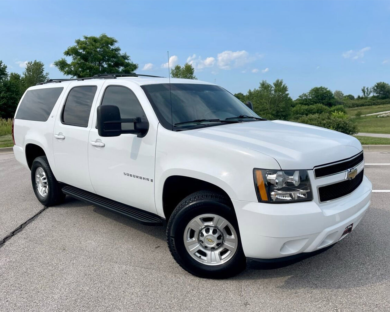 White Chevrolet Suburban 2500 parked with its wheels turned to the left, blue sky with clouds and trees behind the car
