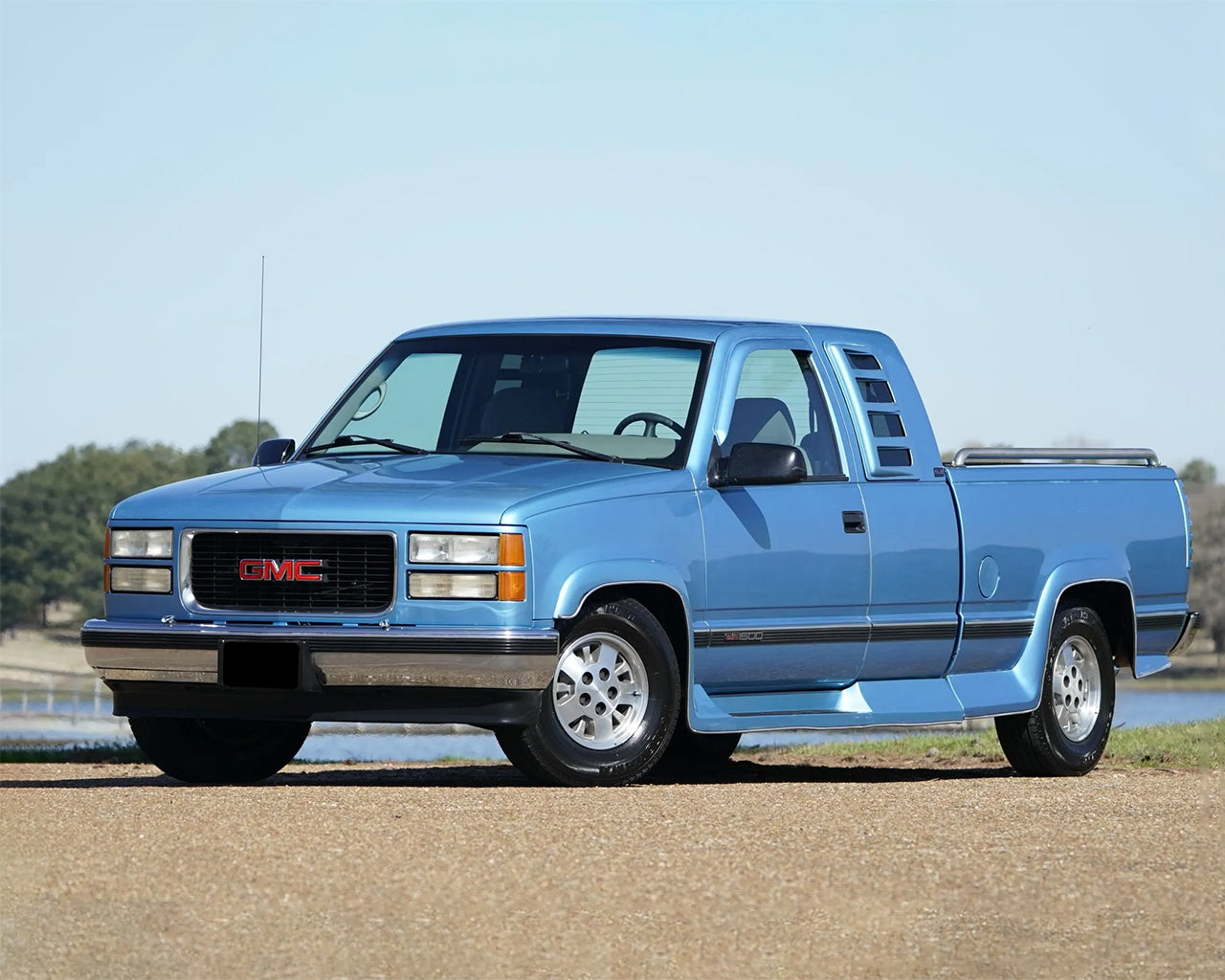 Blue GMC C1500 truck with vents on the side window parked on a road with a lake in the background