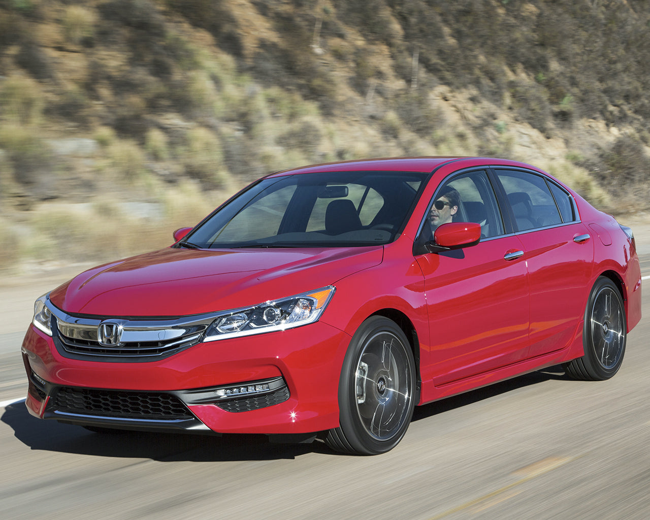 Red Honda Accord driving on a road passing a cliff side covered in small shrubs
