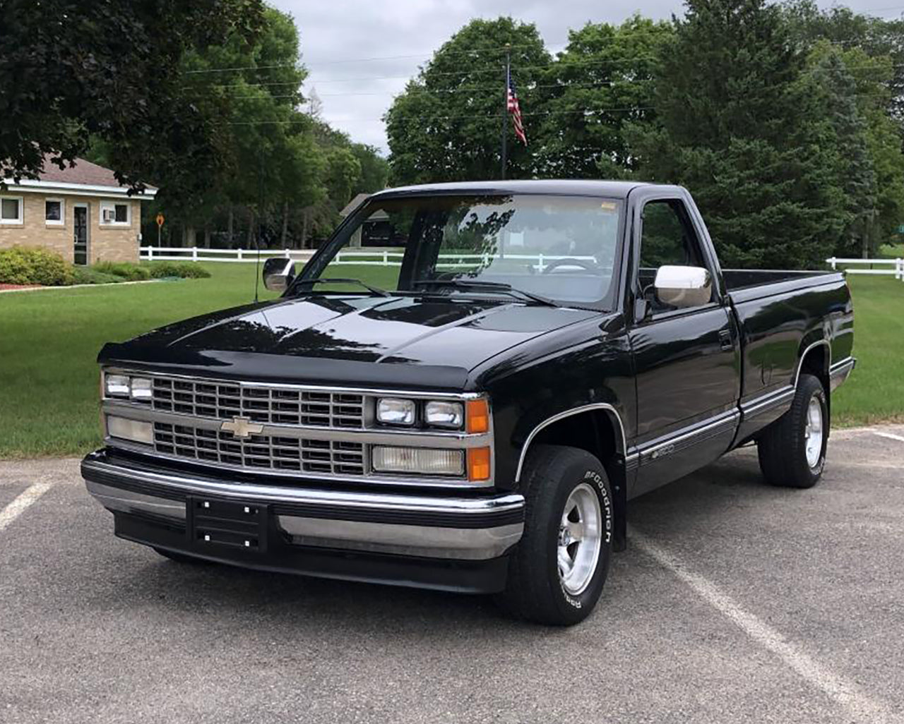Black Chevrolet C1500 parked infront of a grassy yard with a flagpole in front of some trees