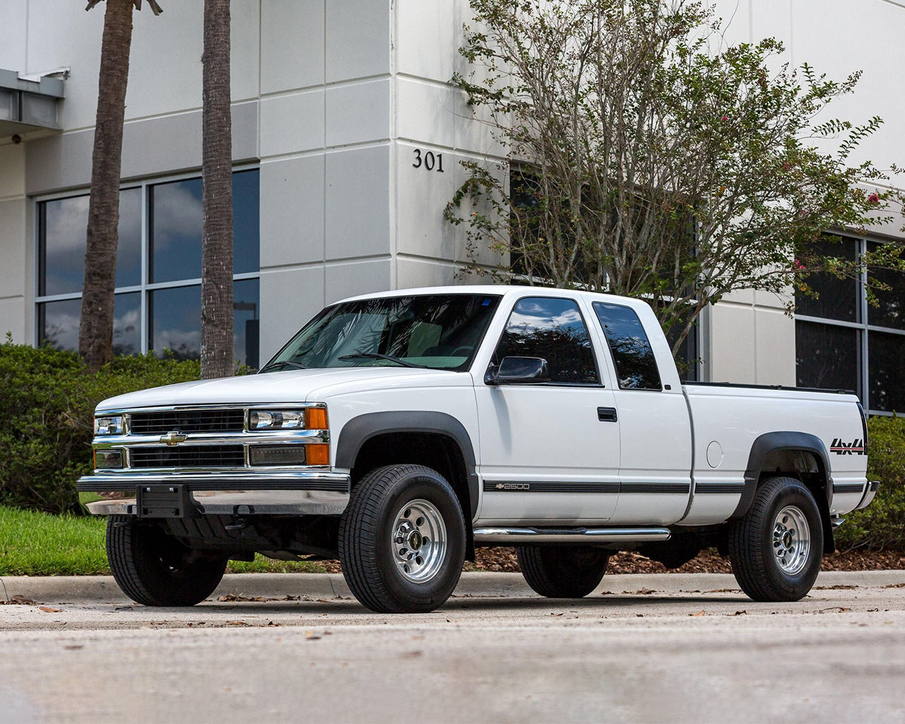 White Chevy K2500 truck parked next to a building with palm trees and grass