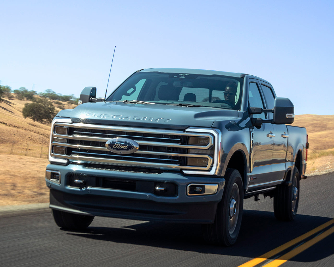 Light blue Ford Super Duty F-350 driving on a road passing grassy hills