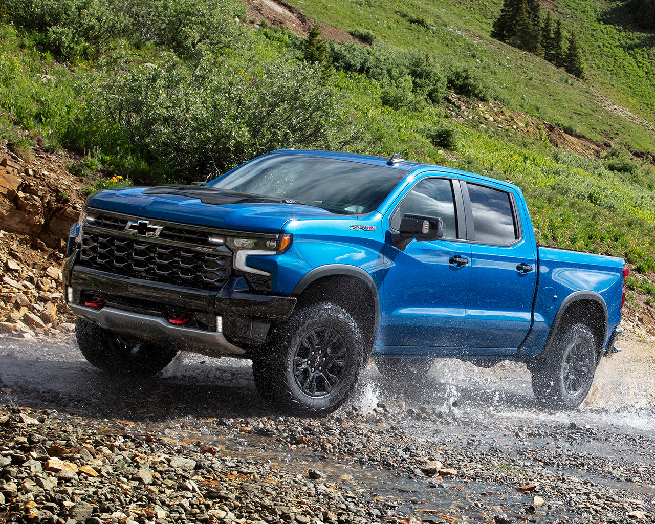Blue Chevrolet Silverado driving through a stream of water and rocks with a grassy hill behind it
