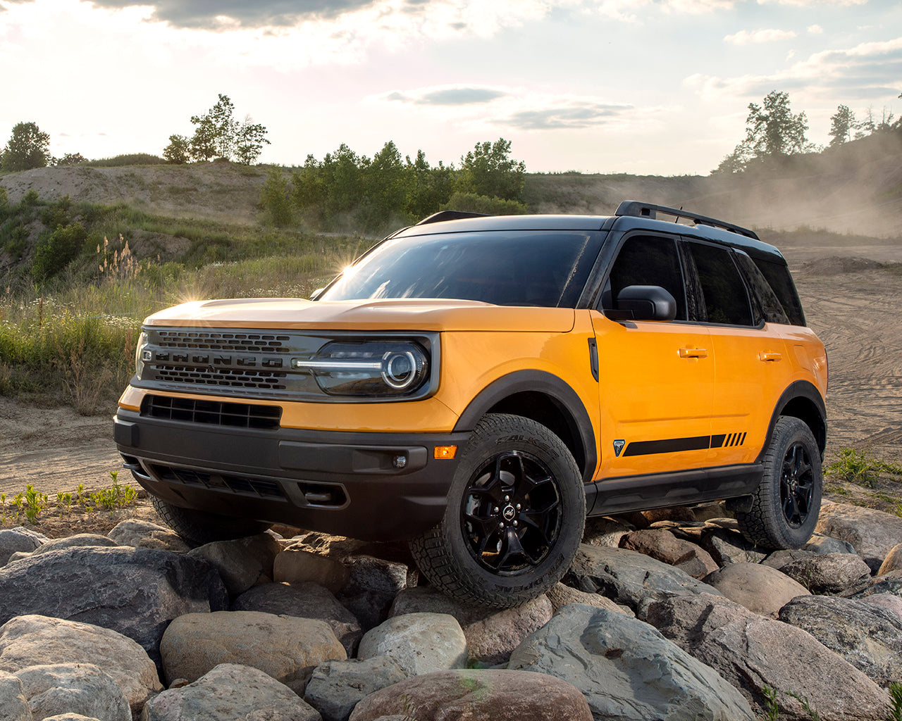 Yellow Bronco Sport parked on a mound of rocks with bushes, trees, and dust clouds behind it