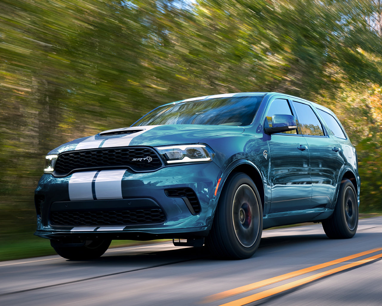 Light Blue Dodge Durango with white racing stripes on the front bumper and hood, driving on a road passing trees in background