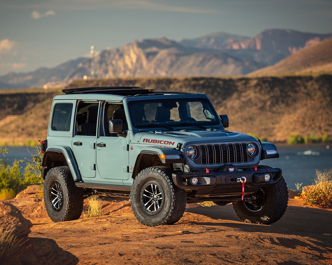 Blue Jeep Wrangler parked on an orange cliff overlooking a lake and mountains